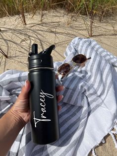 a person holding a black water bottle on top of a towel next to the beach