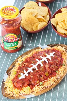 a hot dog covered in chili and topped with football shaped toppings next to chips