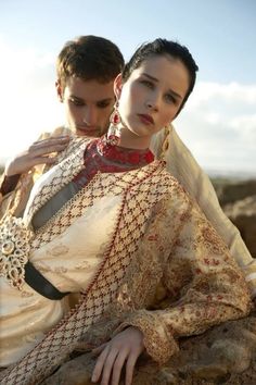 a man and woman sitting next to each other on top of a rocky hill with the sun behind them