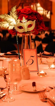 the table is set with glasses, silverware and red roses in a tall vase