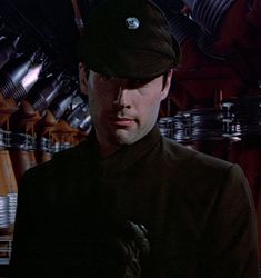 a man wearing a black uniform and hat in a room full of metal pipes, looking at the camera