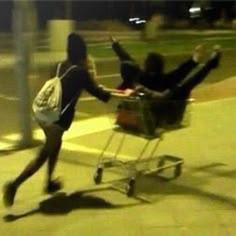 two people pushing a shopping cart down the street at night time with one person on it