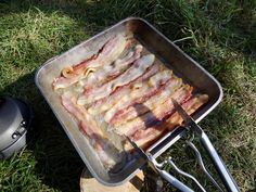 bacon cooking in a pan on the grass with tongs and an oven mitt next to it