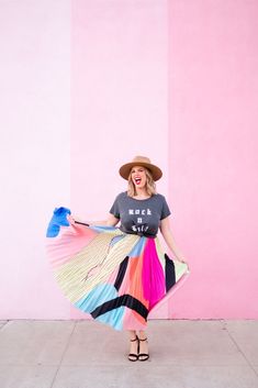 a woman standing in front of a pink wall wearing a colorful skirt and a hat