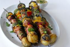 a white plate topped with meat and veggies on skewers next to dipping sauce