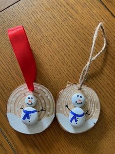 two snowman ornaments hanging from a red ribbon on a wooden table with wood planks