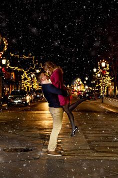 a man and woman kissing in the middle of a street at night with snow falling all around them