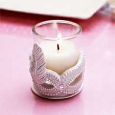 a candle that is sitting in a glass vase on a pink tablecloth with a white plate behind it