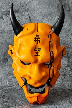 an orange mask with black horns and writing on the face is sitting on a gray surface