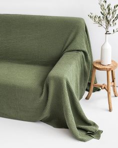 a couch covered with a green blanket next to a small wooden table and white vase