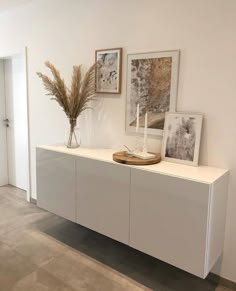 a white cabinet with two pictures on the wall and a vase filled with dried grass