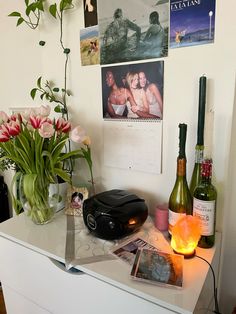 a white dresser topped with bottles and vases filled with flowers next to pictures on the wall