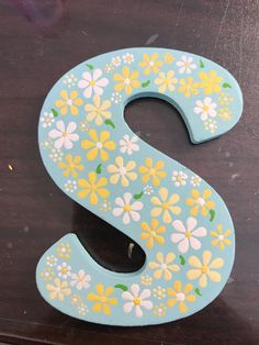 the letter s is painted with flowers and daisies on blue paint, sitting on a wooden surface