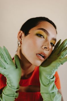 a woman with green gloves and yellow eyeshadow holding her hands up to her face