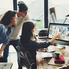 three people sitting at a desk looking at a computer screen with images on it and one person pointing to the monitor