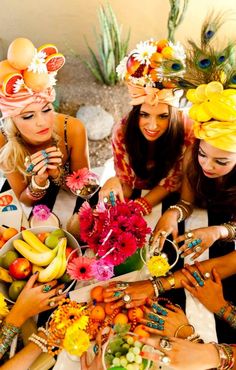 four women wearing headdress around a table with fruit and flowers in their hands