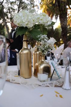 white flowers in gold vases on a table with other decorations and people standing around