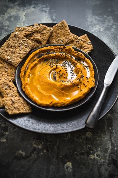 a black plate topped with crackers and an orange hummusal dip surrounded by crackers