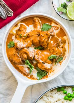 two bowls filled with chicken curry and rice next to a plate of peas, lime wedges and cilantro