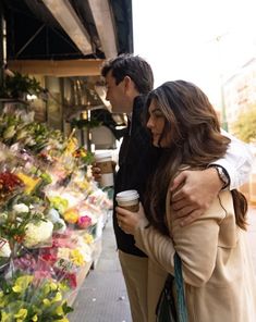 two people standing next to each other in front of flowers and coffee cups on the sidewalk