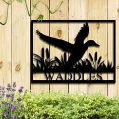 a metal sign that says waddles with a bird flying over it in front of some flowers