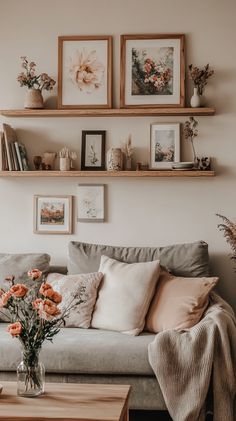 Cozy living room with gray sofa, pastel pillows, wooden shelves, framed art, and warm lighting, showcasing a boho aesthetic. Floating Shelves Living Room Above Couch, Living Room Decor Over Couch, Wall Picture Collage Ideas Living Rooms, Living Room Designs Cozy Boho, Earthy Cozy Living Room, Cosy Neutral Living Room, Cozy Couch Aesthetic, Above Couch Wall Decor Farmhouse, Above Couch Wall Decor Boho