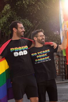 two men standing next to each other in front of a rainbow flag and one holding a gay pride t - shirt