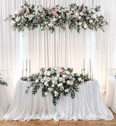 an arrangement of flowers and greenery on a white table cloth with candles in the background