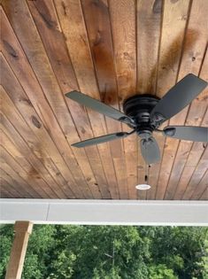 a ceiling fan mounted to the side of a wooden porch with trees in the background