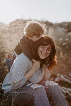 a woman holding a young boy in her arms while sitting on top of a field