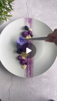 a white plate topped with purple flowers and berries next to a person holding a knife