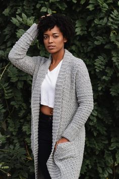 a woman standing in front of a bush wearing a gray cardigan