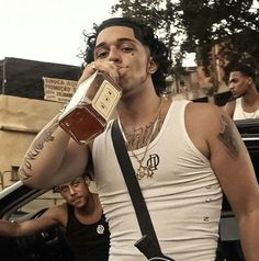 a man drinking from a bottle while standing next to a car with other men in the background