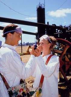 two people standing next to each other in front of an old machinery wheel and building