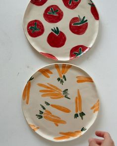 two plates with tomatoes and carrots painted on them, one being held up by a person's hand