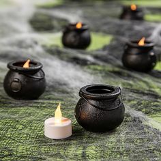 several black pots with lit candles sitting on a green cloth covered tablecloth in front of them