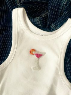 a white tank top with an orange and pink drink embroidered on the front, next to a blue striped shirt