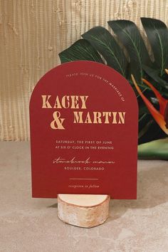 a red and gold wedding card on top of a wooden stand next to a potted plant