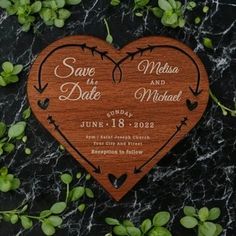 a wooden save the date heart on a black marble surface with greenery around it