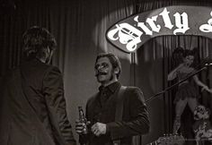 a man in a suit and tie holding a beer next to another man with a guitar