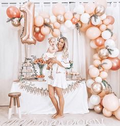 a woman holding a baby standing in front of a cake table with balloons and decorations