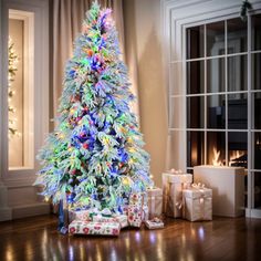 a brightly lit christmas tree with presents on the floor in front of a fireplace and windows