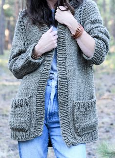 a woman standing in the woods with her hands on her chest and wearing a green knitted cardigan