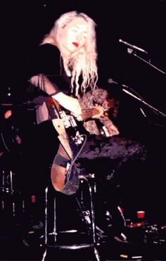 a woman with long white hair sitting on a stool playing an acoustic guitar in front of microphones