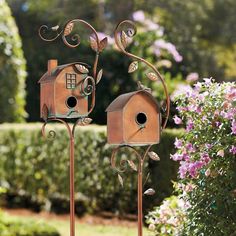 two bird houses on top of metal stands in front of bushes and flowers with purple flowers