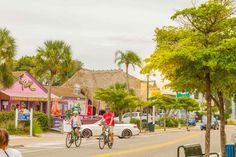 two people riding bikes down the street in front of small shops and palm trees on both sides