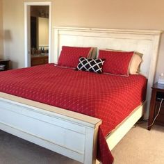 a bed with red sheets and pillows in a bedroom next to a dresser, mirror and lamp