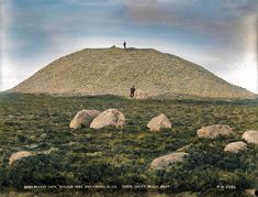 two people standing on top of a hill with rocks in the foreground and green grass below