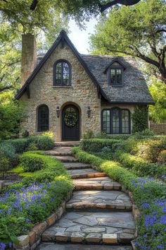 a stone house with steps leading up to the front door and landscaping around it, surrounded by blue flowers