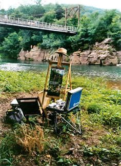 an artist's easel sitting in the grass next to a river under a bridge